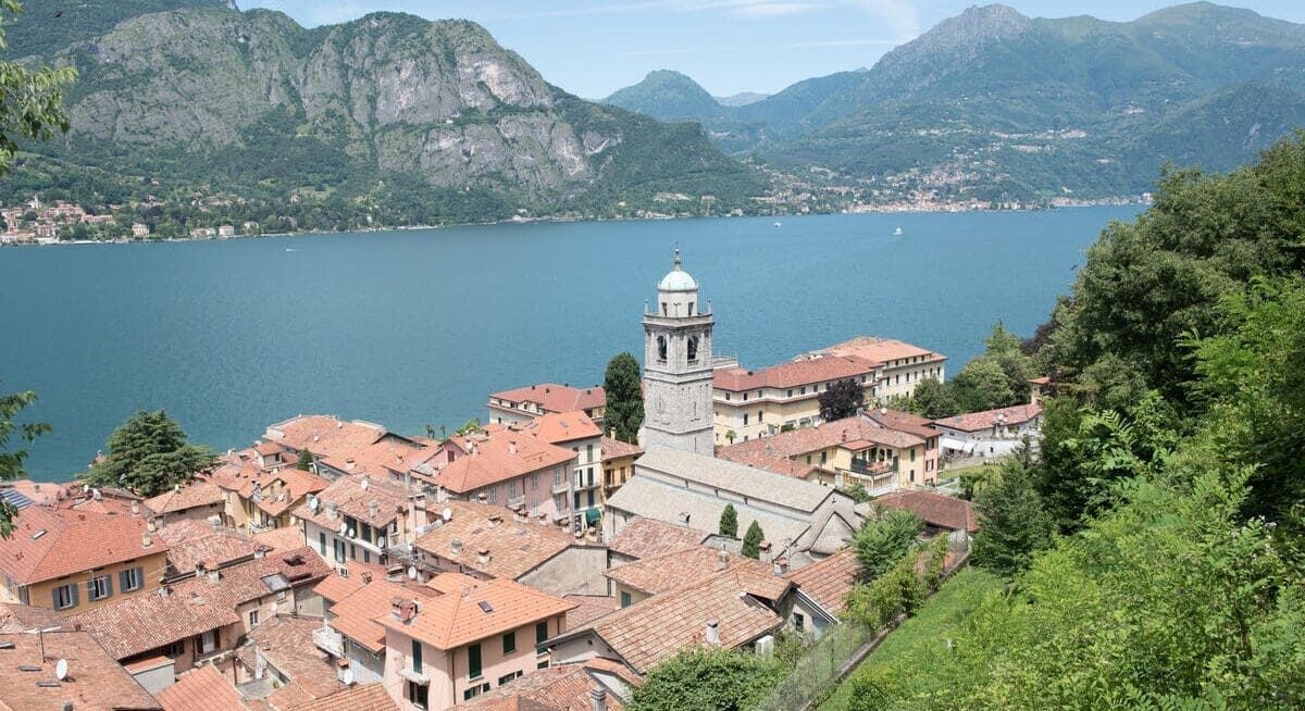 view of a lakeside town with red-roofed buildings and a central church tower, surrounded by green hills and a large blue lake