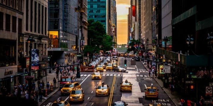 busy city street with numerous yellow taxis and people on sidewalks. Tall buildings line the street, and the sunset is visible in the background